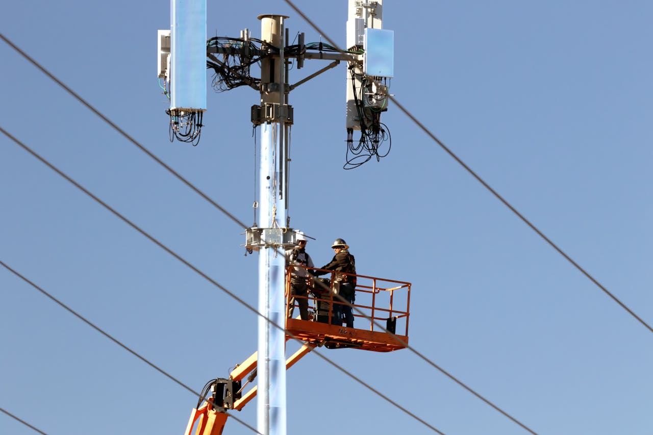 Hakkımızda Technicians on a lift installing communication devices on a utility pole against a clear sky.