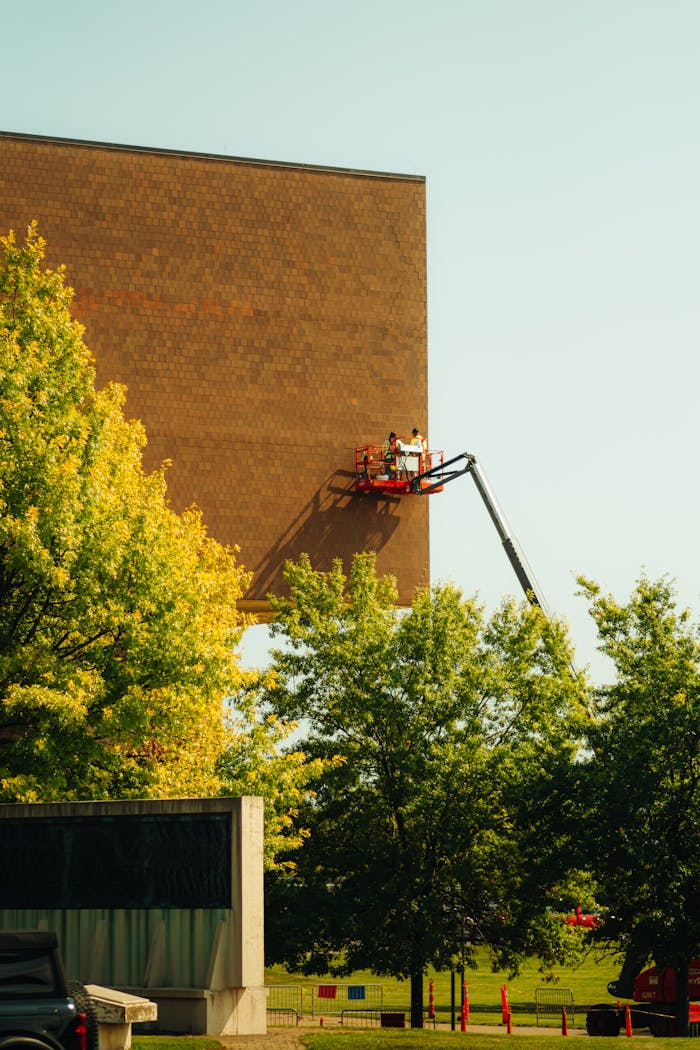 Anasayfa Construction workers repairing a large building facade using a boom lift amidst green trees.
