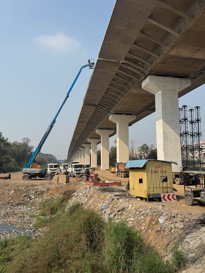 Anasayfa View of elevated roadway construction using a crane and trucks, showcasing infrastructure development in Delhi, India.